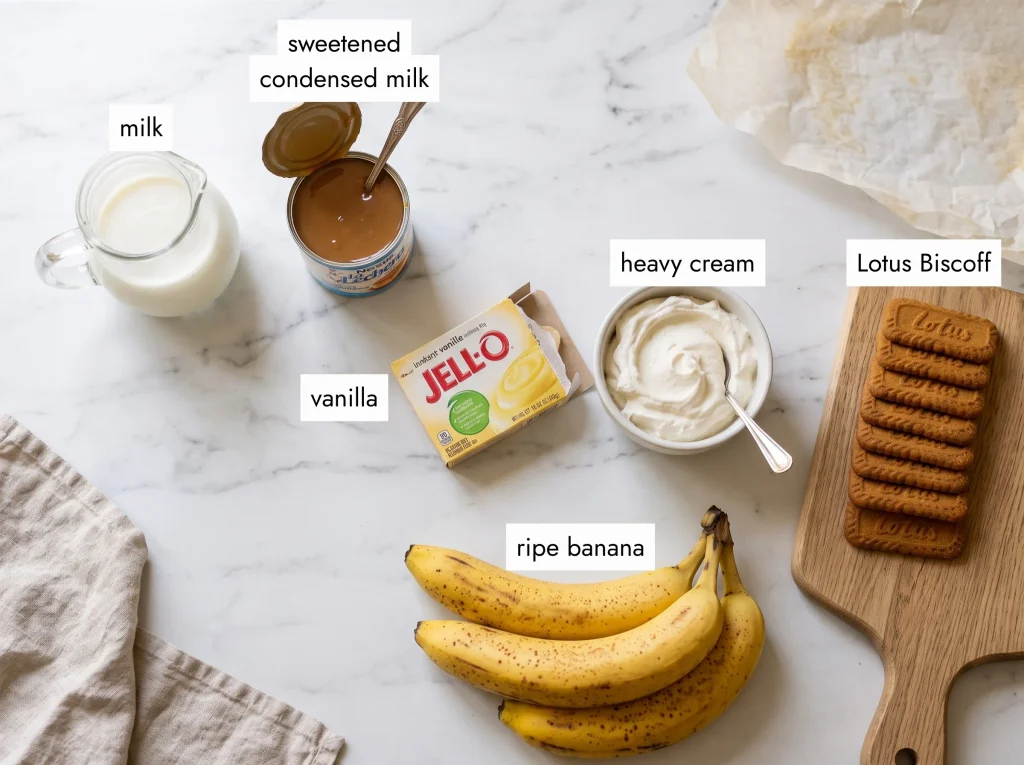 all ingredients for biscoff banana pudding recipe laid out on white marble — lotus biscoff cookies, ripe bananas, condensed milk, vanilla pudding mix and heavy cream