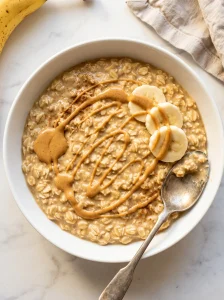 Overhead view of peanut butter banana overnight oats in a white bowl with peanut butter drizzle, fresh banana slices and a vintage spoon