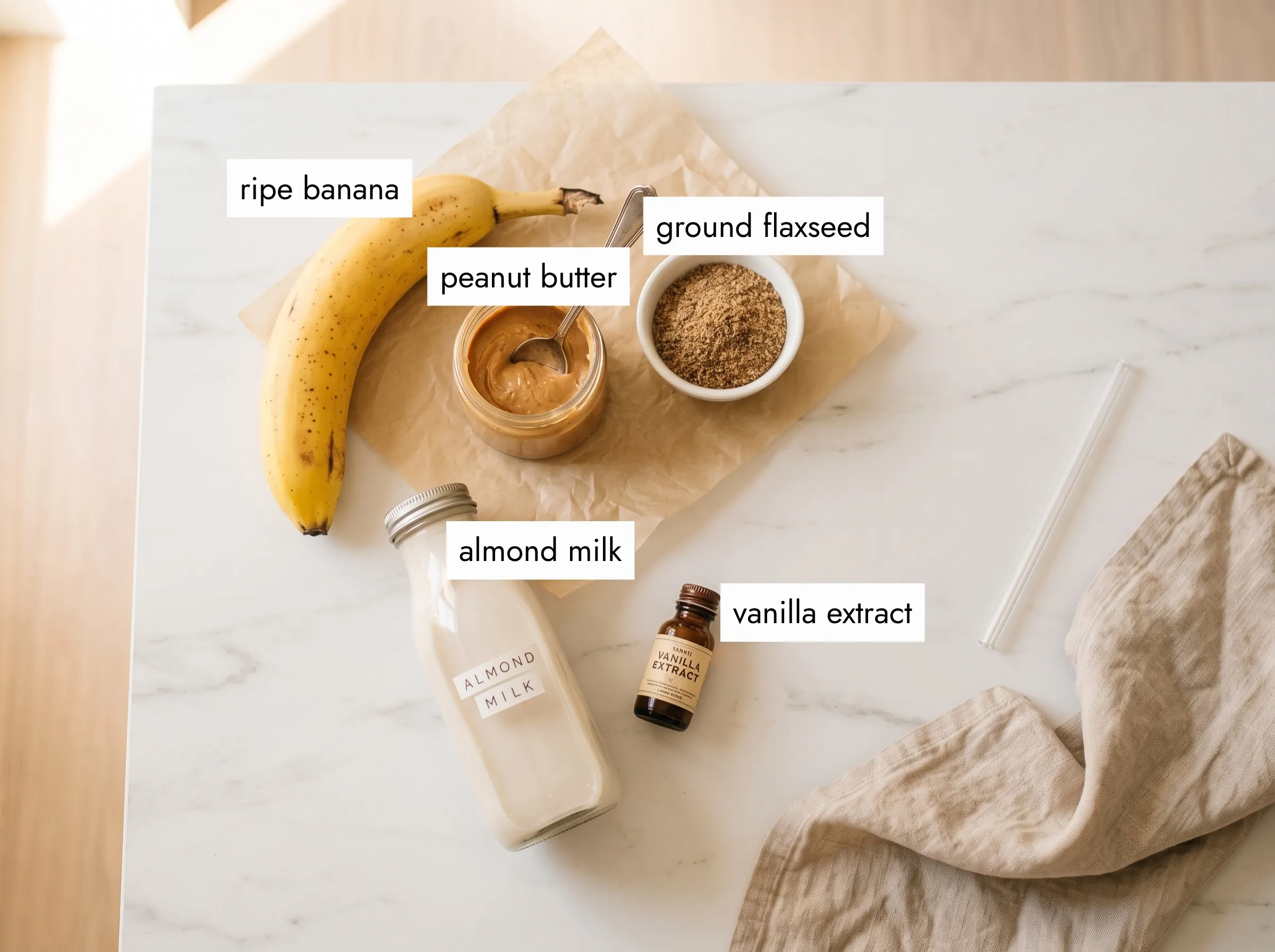overhead flat lay of peanut butter banana smoothie ingredients — frozen banana, natural peanut butter jar, almond milk, ground flaxseed on white marble