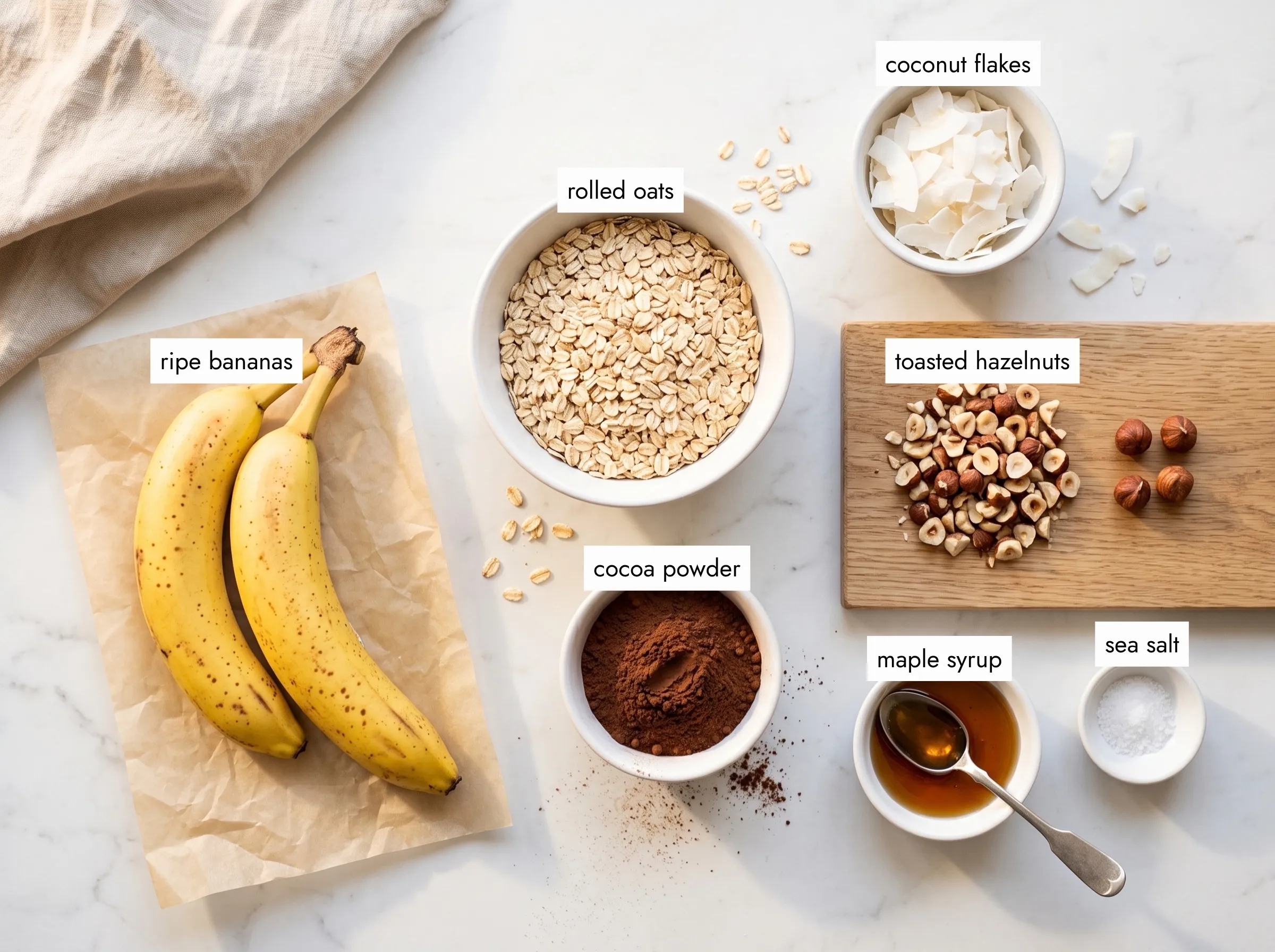 Ingredients for a banana bites recipe: ripe bananas, rolled oats, cocoa powder, chopped hazelnuts, coconut flakes and maple syrup