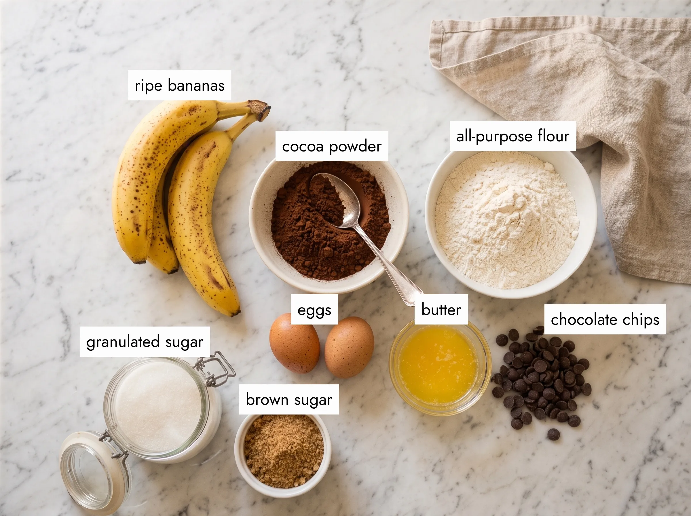 Overhead flat lay of chocolate banana cake ingredients on white marble — ripe bananas, cocoa powder, flour, eggs, butter, chocolate chips