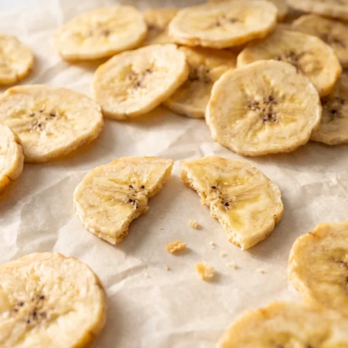 Close-up of crispy dehydrated banana chips on crumpled parchment, one snapped in half to show its dry, wafer-thin texture