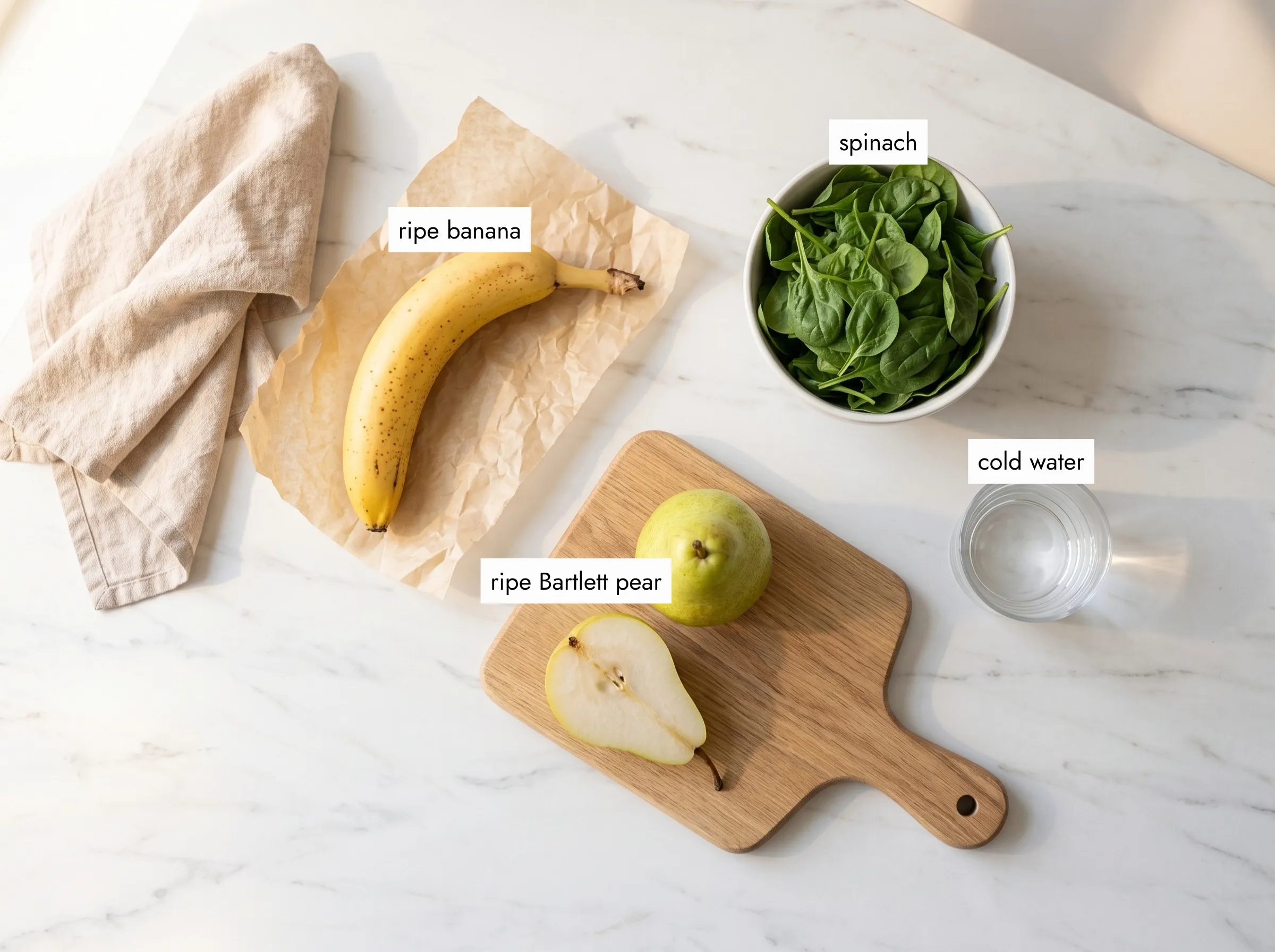 Four green banana smoothie ingredients on white marble: ripe banana, Bartlett pear, baby spinach and cold water