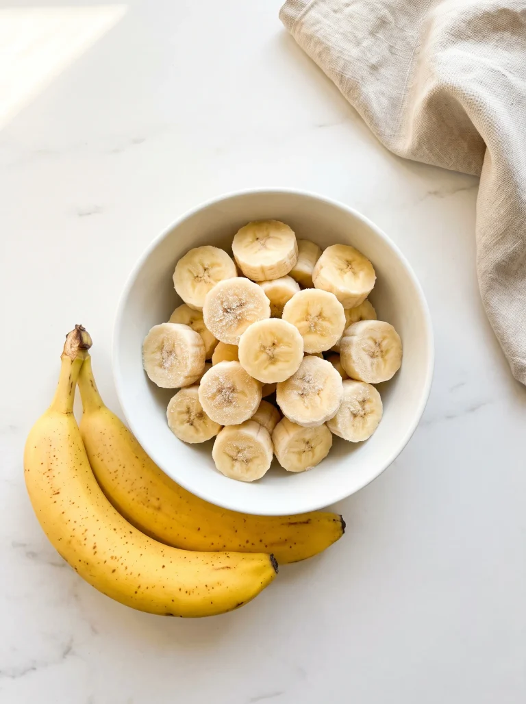White bowl of frozen banana slices with two ripe golden bananas on white marble, how to freeze bananas